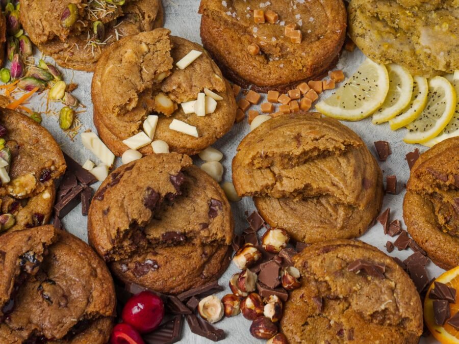The full cookie range photographed overhead as a wide banner, each flavour styled with its own ingredients including grated carrot, pistachio, cranberry, caramel, lemon, hazelnut and orange, artisan cookie photography in Croydon.