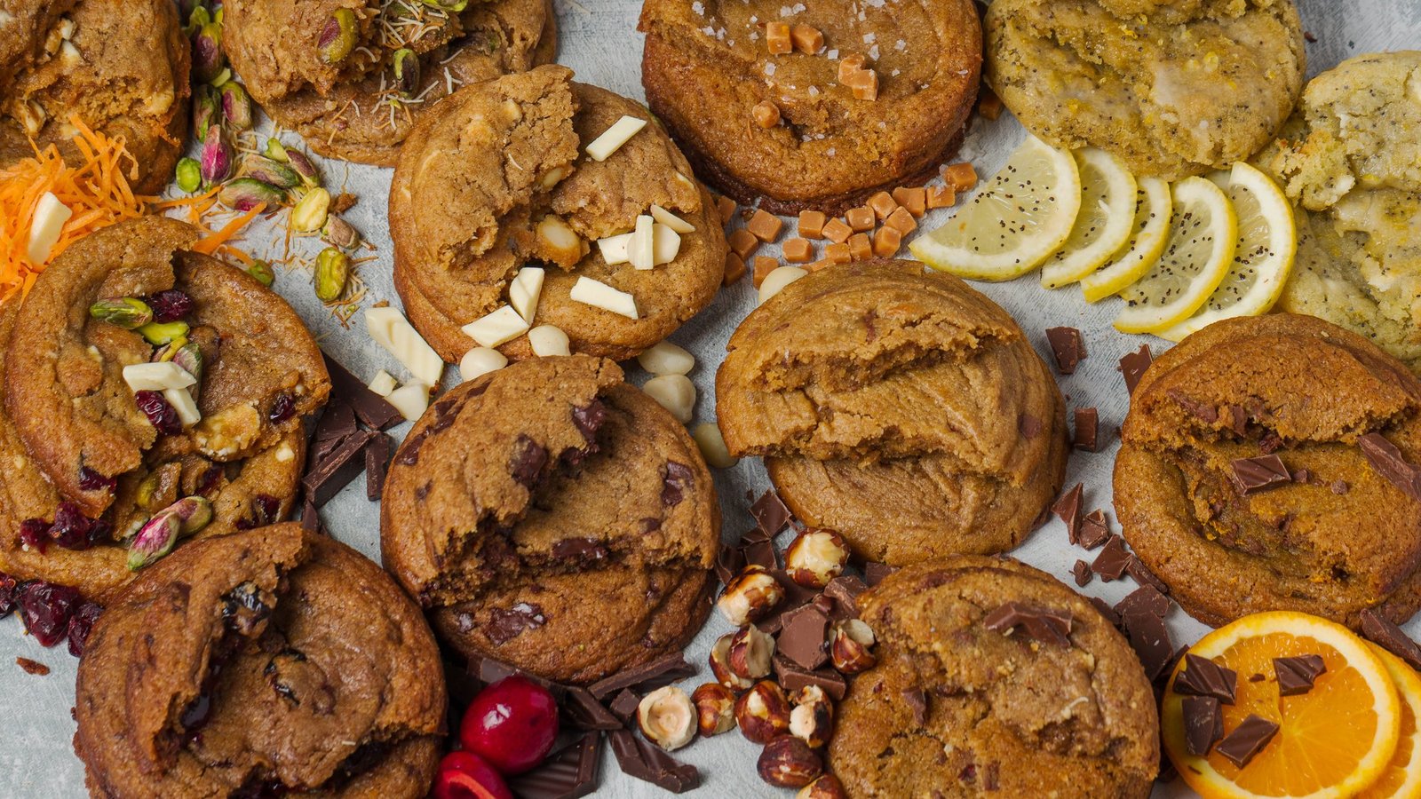 The full cookie range photographed overhead as a wide banner, each flavour styled with its own ingredients including grated carrot, pistachio, cranberry, caramel, lemon, hazelnut and orange, artisan cookie photography in Croydon.