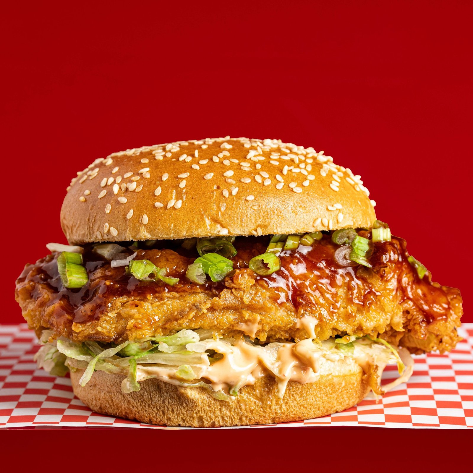 A fried chicken burger under a glossy glaze with sliced spring onion on a sesame bun with dressed shreds, delivery app photography for a halal chicken shop in Wood Green.