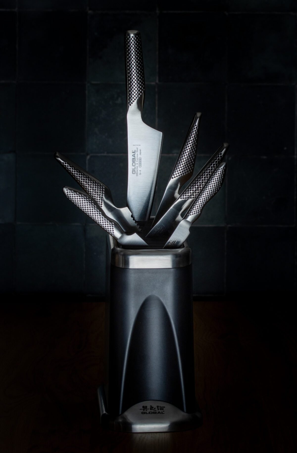 Global knives fanned out of a dark knife block against black tiles, lit low and moody