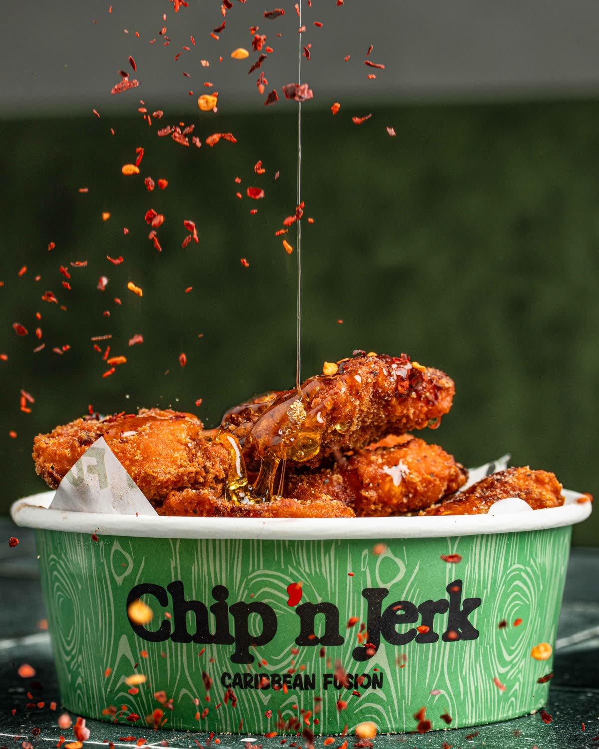 Honey pouring over fried chicken in a branded tub with chilli flakes falling through the frame, motion food photography for a Caribbean fusion QSR in Holloway, London.