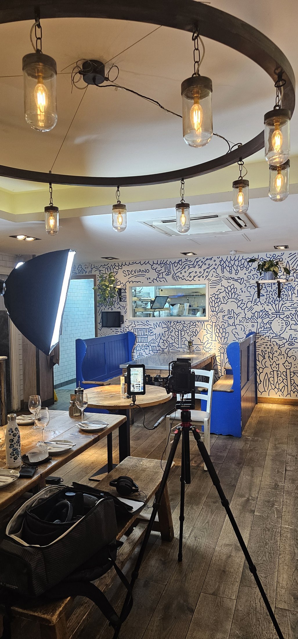 An interview lit and framed inside The Real Greek in Soho, softbox and tripod set against the restaurant's blue and white mural, behind the scenes of a London restaurant video production.
