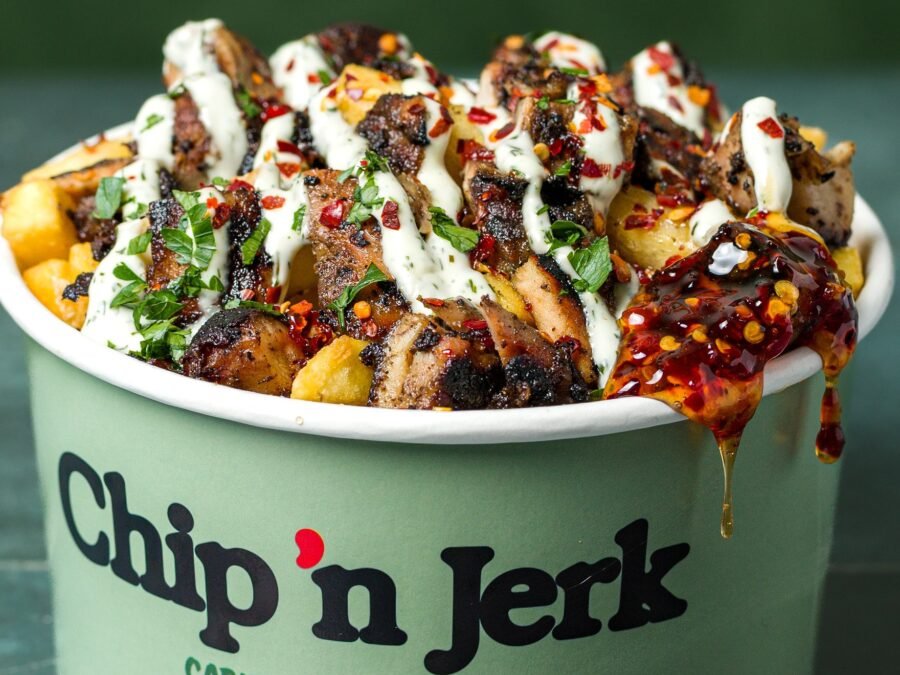 Jerk chicken loaded fries in a branded Chip 'n Jerk tub with herb sauce, chilli flakes and a scotch bonnet sauce drip running over the rim, food photography for a Caribbean QSR in Holloway, London.