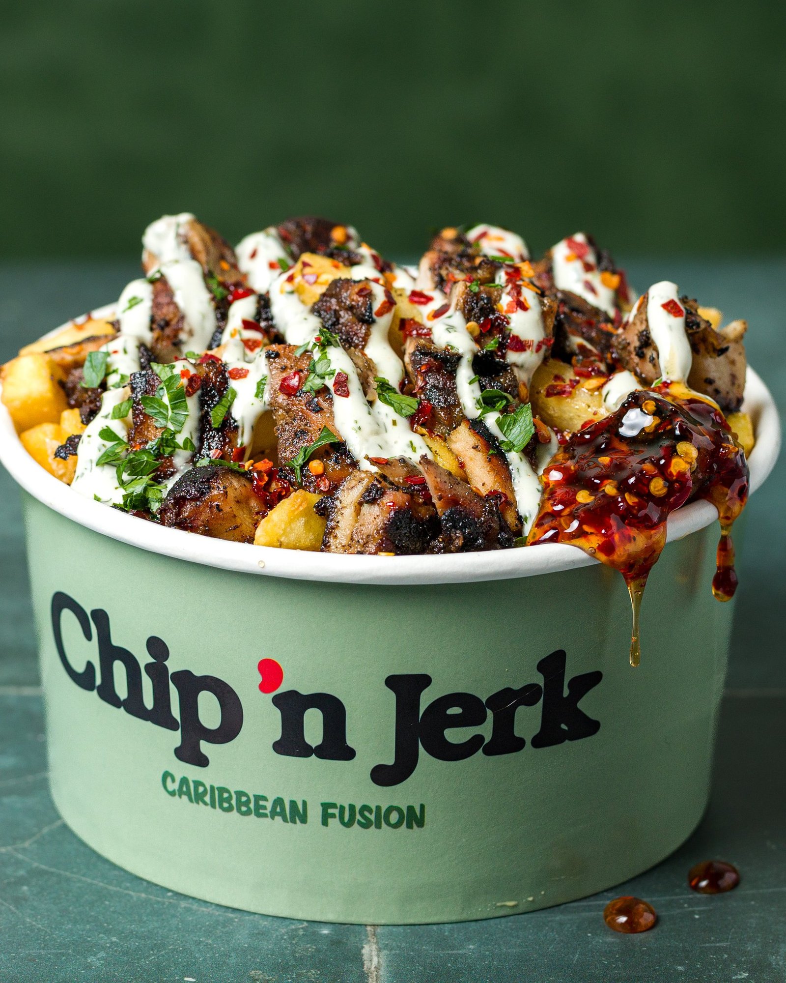 Jerk chicken loaded fries in a branded Chip 'n Jerk tub with herb sauce, chilli flakes and a scotch bonnet sauce drip running over the rim, food photography for a Caribbean QSR in Holloway, London.