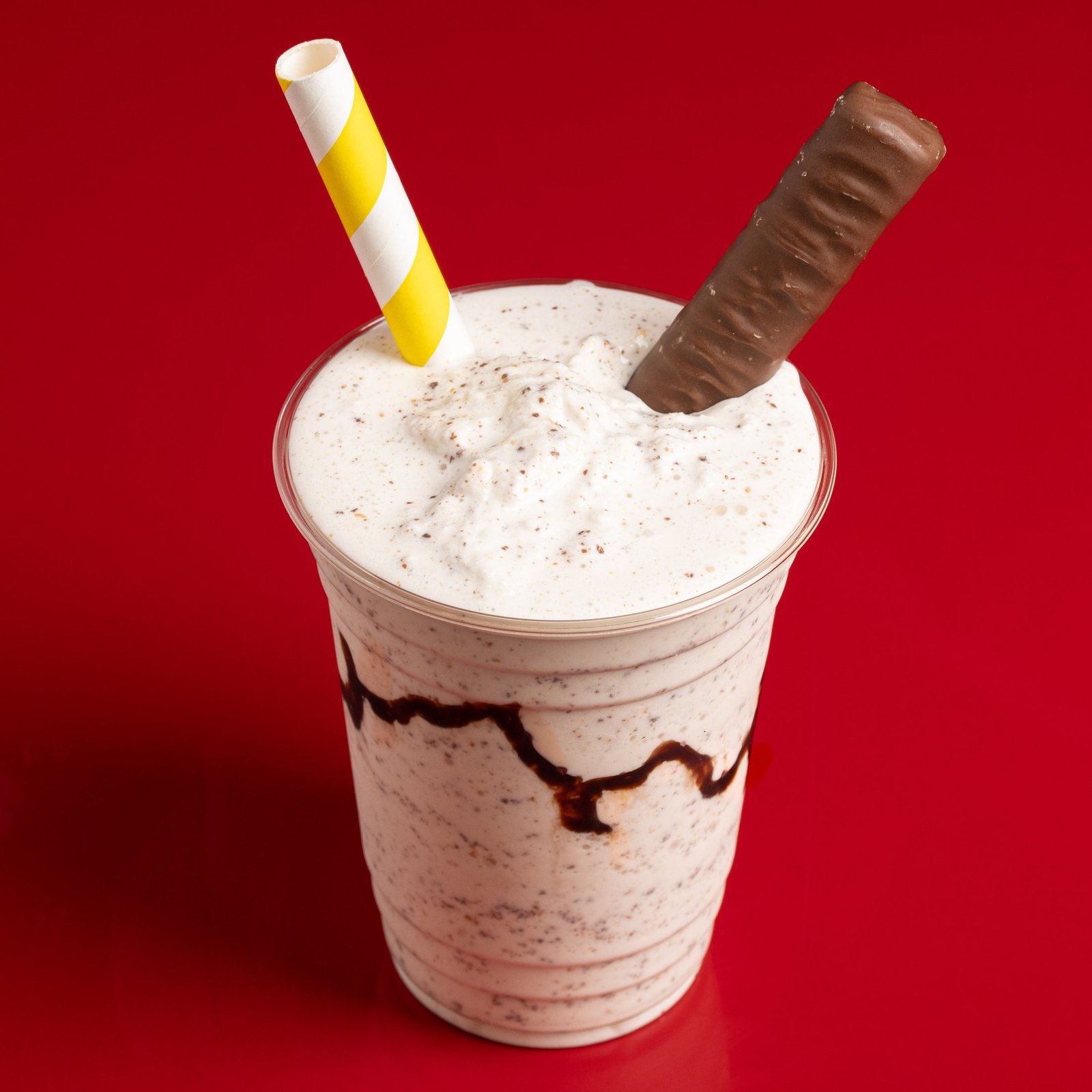 A shake in a clear takeaway cup with a whipped top, chocolate sauce running down the inside of the cup, a chocolate bar and a striped paper straw, menu photography for a chicken shop in Wood Green.