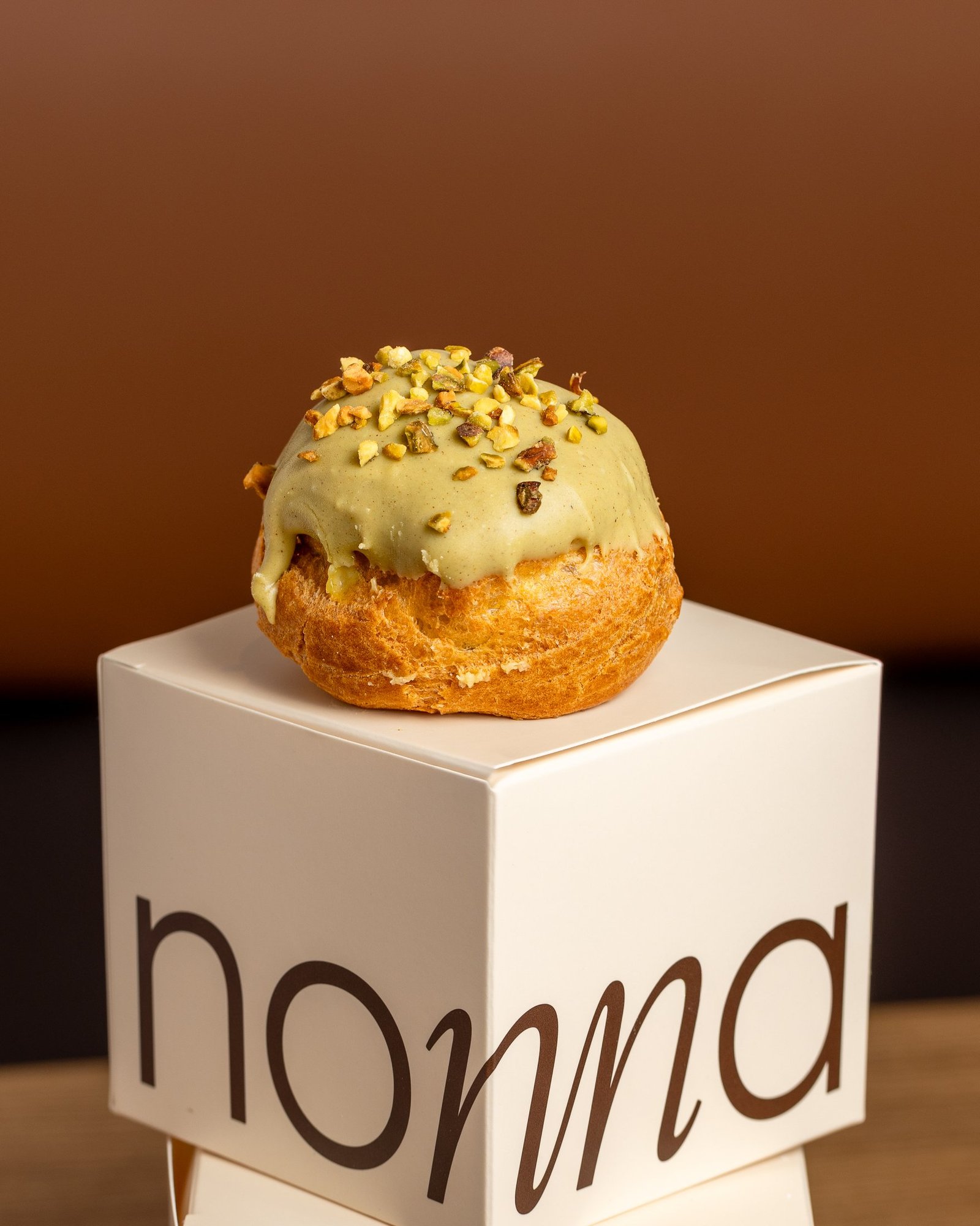 Pistachio bignè, a choux bun under pistachio glaze scattered with chopped pistachios, standing on a branded nonna box, pastry photography for a Sicilian bakery in Holborn.