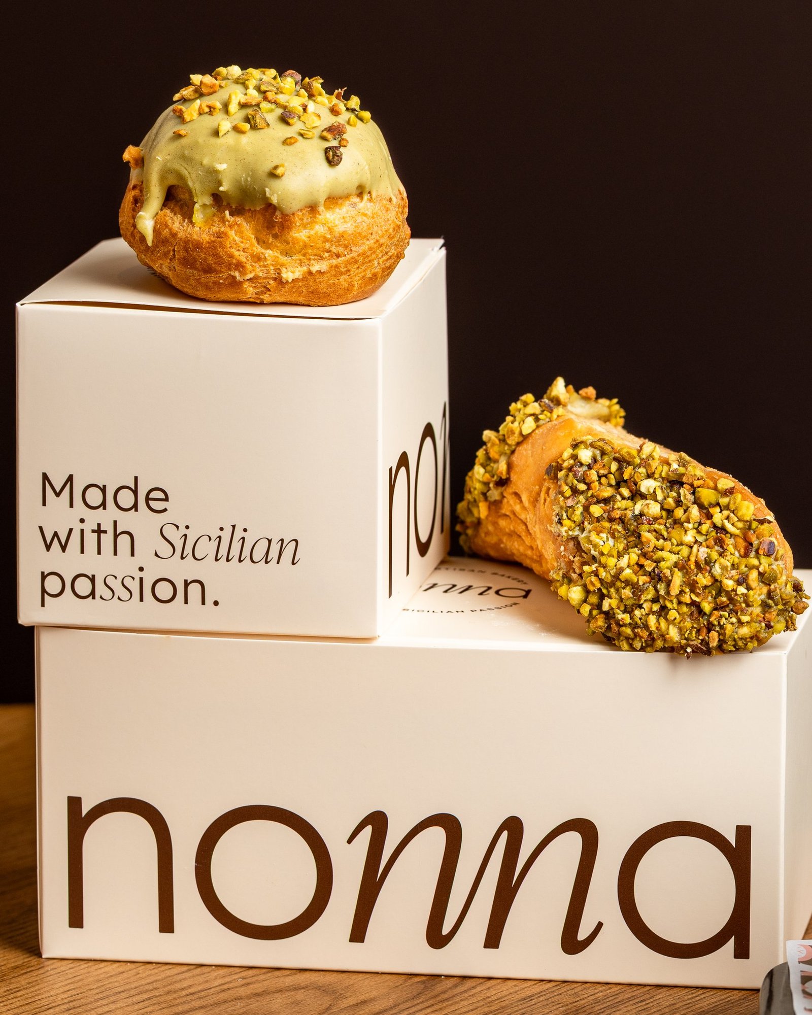 A pistachio cannolo rolled in chopped pistachio beside a glazed pistachio bignè, staged on branded nonna boxes reading Made with Sicilian passion, bakery photography in Holborn, London.