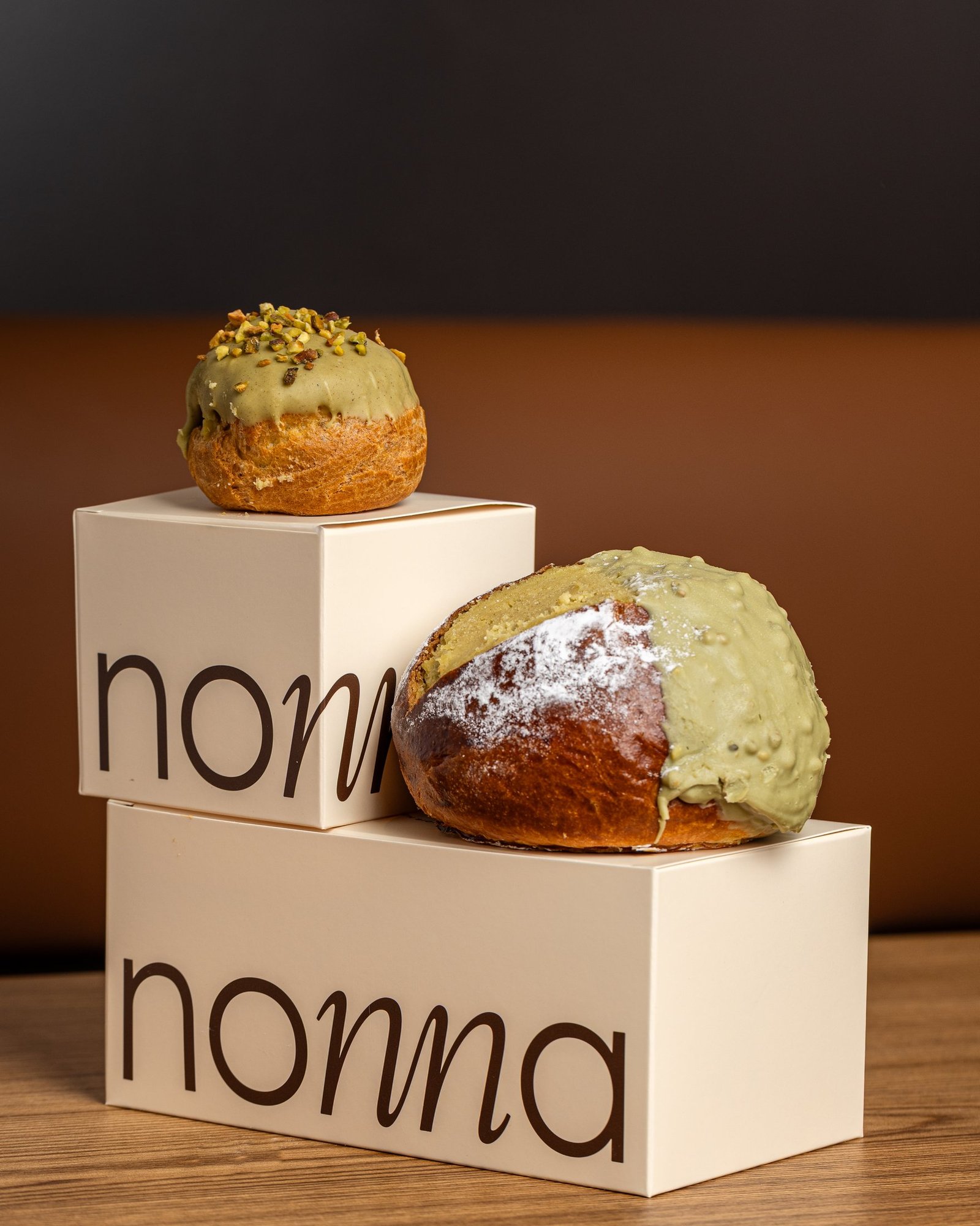 A pistachio maritozzo, brioche split and filled with cream and half dipped in pistachio, beside a pistachio bignè on branded nonna boxes, Sicilian pastry photography in Holborn.
