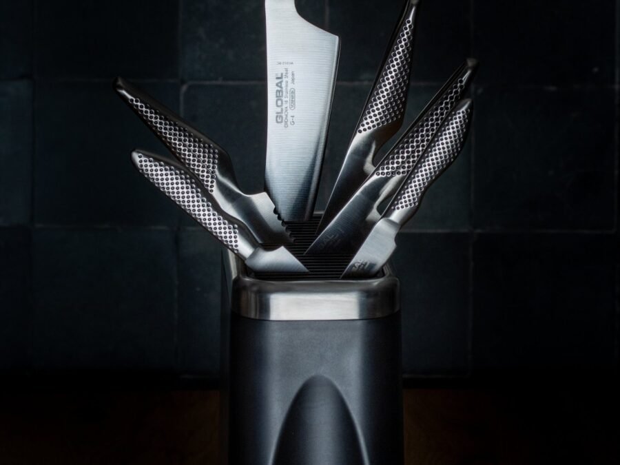 A Global knife block with a set of premium Japanese knives fanned out of it, lit against dark teal tiles, product photography for a premium kitchen knife brand.