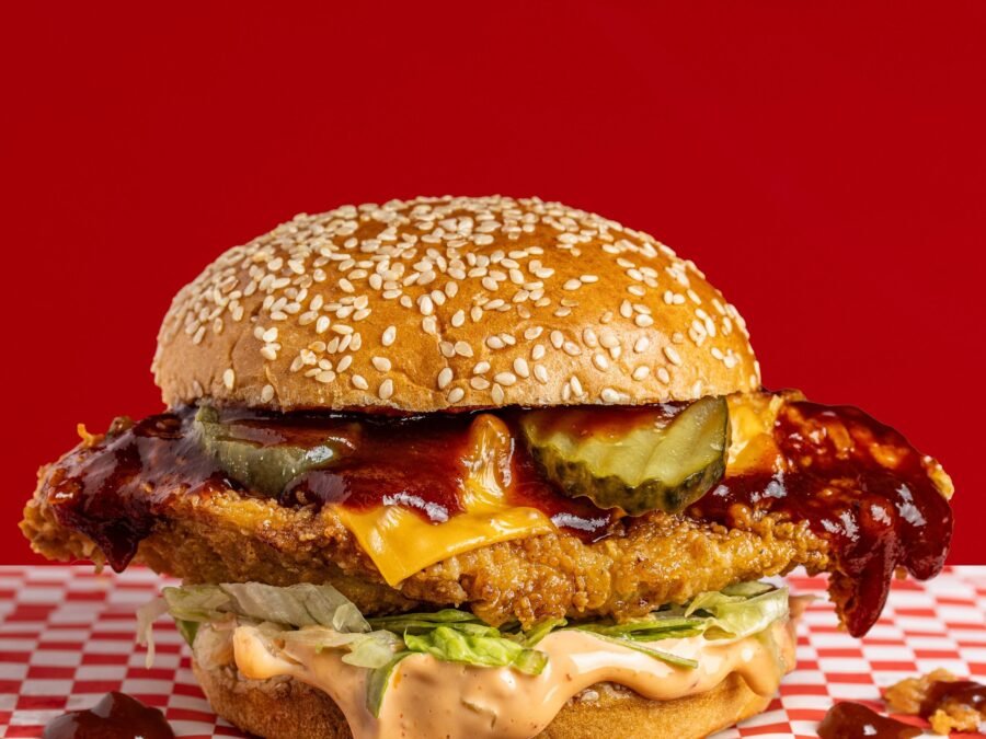 A fried chicken burger with a dark glossy sauce, melted cheese and pickles, burger sauce spilling onto checked paper, food styling and menu photography for a chicken shop in Wood Green.