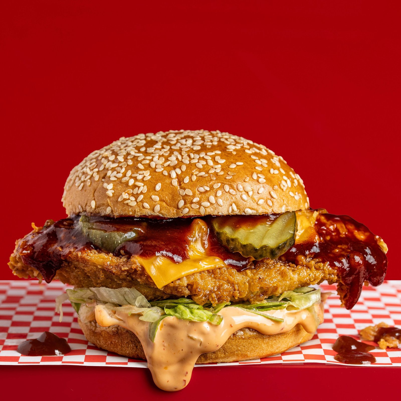 A fried chicken burger with a dark glossy sauce, melted cheese and pickles, burger sauce spilling onto checked paper, food styling and menu photography for a chicken shop in Wood Green.